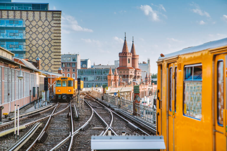 Blick auf die U-Bahn-Gleise in Berlin mit einem gelben Zug im Vordergrund und der markanten Oberbaumbrücke im Hintergrund, umgeben von modernen und historischen Gebäuden.