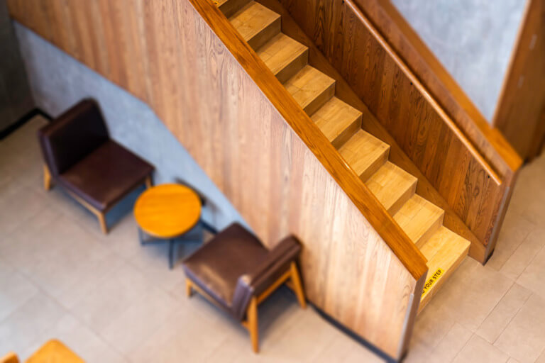 Holztreppe in modernem Innenraum mit gemütlichen Sitzgelegenheiten und Tischen. Die Treppe führt zu einem oberen Bereich und ist Teil eines stilvollen, minimalistischen Designs.