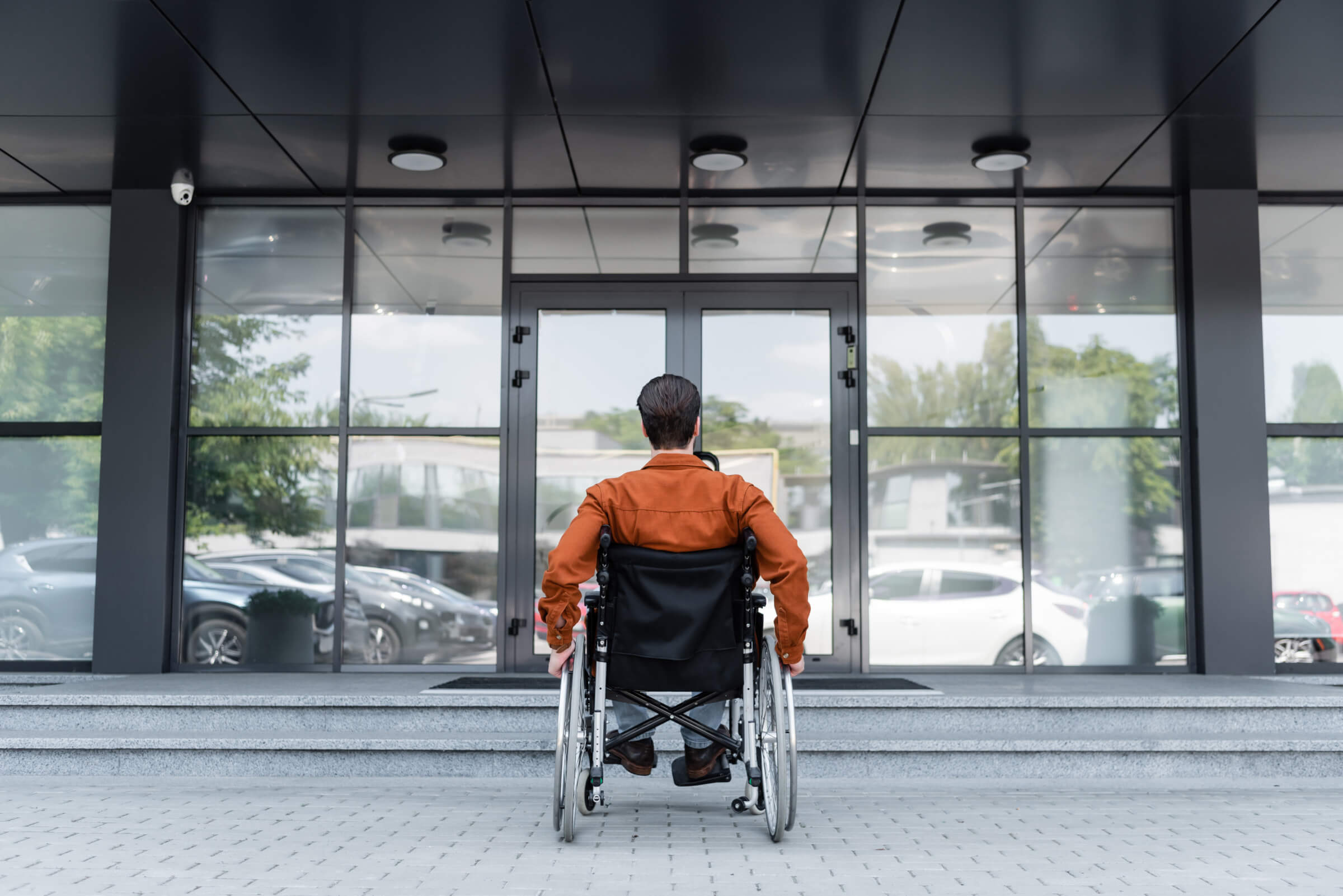Mann im Rollstuhl vor einem modernen Gebäude mit großen Glasfenstern, der auf den Eingang blickt, symbolisiert Barrierefreiheit und Inklusion.