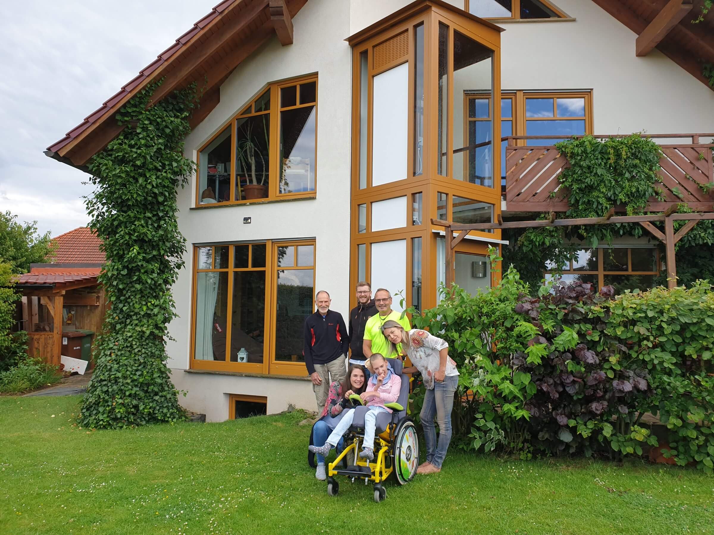 Familie posiert fröhlich im Garten vor einem modernen, lichtdurchfluteten Haus mit großen Fenstern und grüner Bepflanzung. Eine Person sitzt im Rollstuhl, während die anderen stehend oder kniend um sie herum stehen und lächeln.