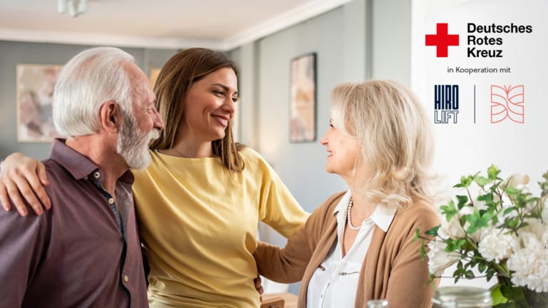 Ein fröhliches Treffen zwischen drei Menschen in einem hellen Raum, das die Verbundenheit und Unterstützung durch das Deutsche Rote Kreuz symbolisiert. Die Szene zeigt eine junge Frau, die liebevoll mit einem älteren Mann und einer älteren Frau spricht, während im Hintergrund Dekoration und Pflanzen für eine einladende Atmosphäre sorgen.