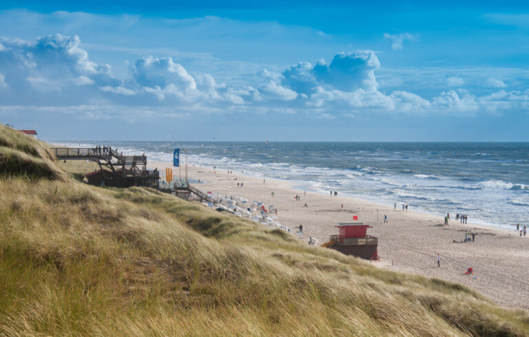 Blick auf einen breiten Sandstrand mit leicht aufgewühltem Meer und vielen Besuchern, umgeben von sanften Dünen unter einem strahlend blauen Himmel mit ein paar Wolken. Ideal für Strandurlaub und Outdoor-Aktivitäten.