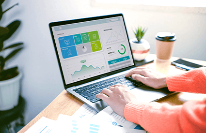 Person arbeitet an einem Laptop, der eine Dashboard-Ansicht mit Grafiken und Statistiken zeigt, während auf dem Tisch Unterlagen und eine Kaffeetasse stehen. Im Hintergrund ist eine Pflanze sichtbar.