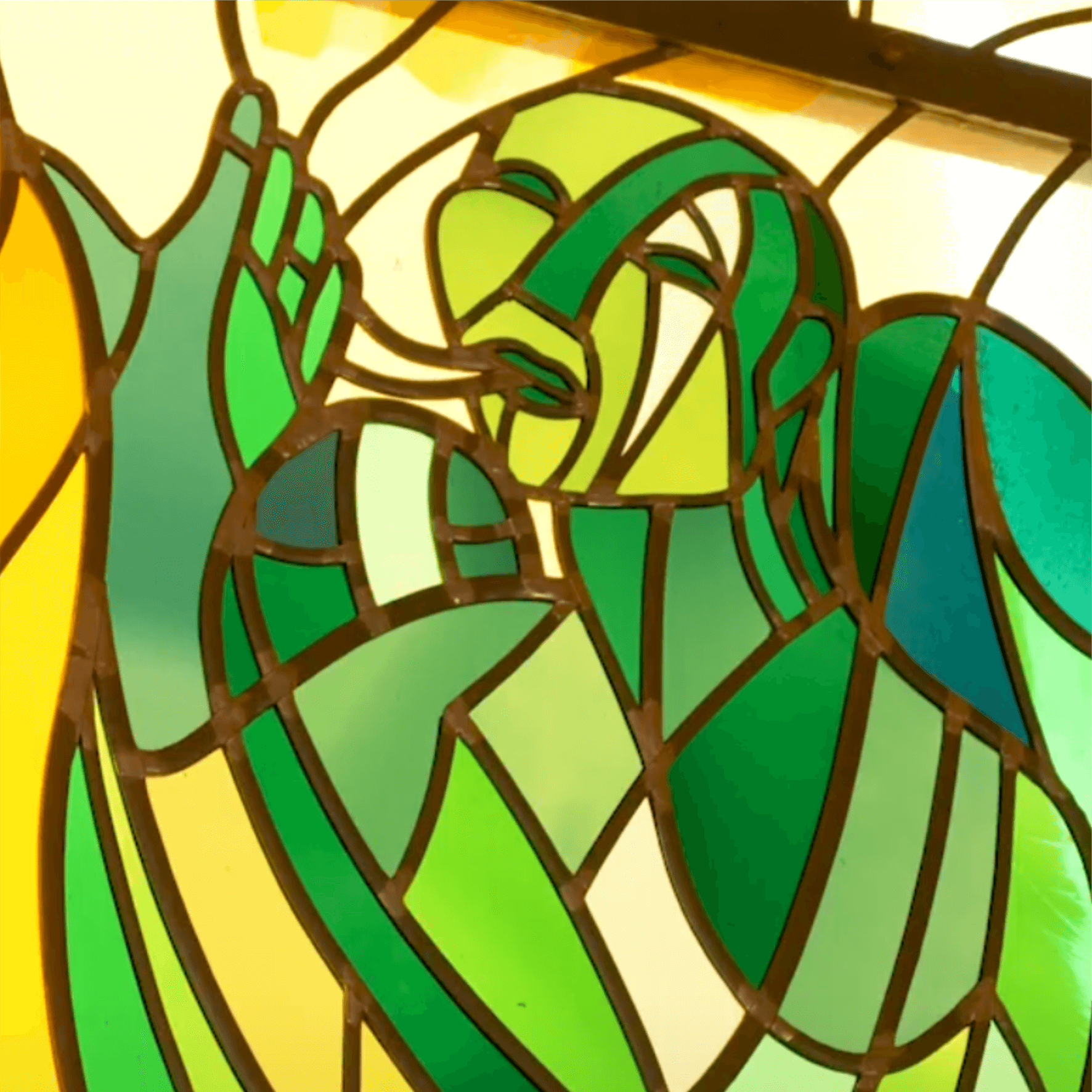 Buntes Glasfenster mit einer stilisierten Figur, die in einem kräftigen Grünton dargestellt ist und eine Handbewegung macht, die eine emotionale Ausdrucksform symbolisiert. Die geometrischen Formen und Farben schaffen eine harmonische Komposition.