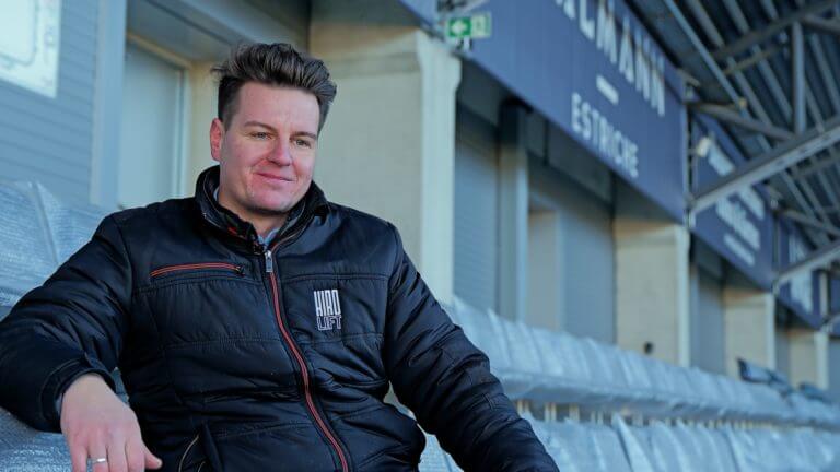 Mann sitzt entspannt auf einer Tribüne im Stadion und lächelt, im Hintergrund sind leere Sitzreihen und ein Schild mit dem Vereinsnamen zu sehen.