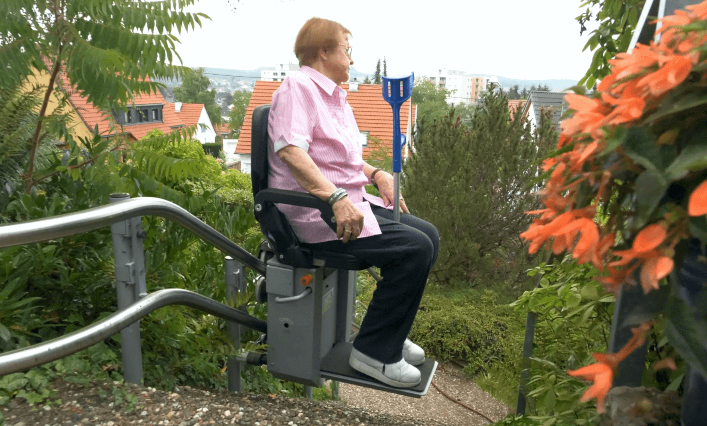 Garten_Weinmann_Treppenlift_Außenanlage_Fahrt Seniorin nutzt eine Treppenliftanlage, um eine steile Gartenstraße zu befahren, umgeben von grünen Pflanzen und blühenden Blumen.