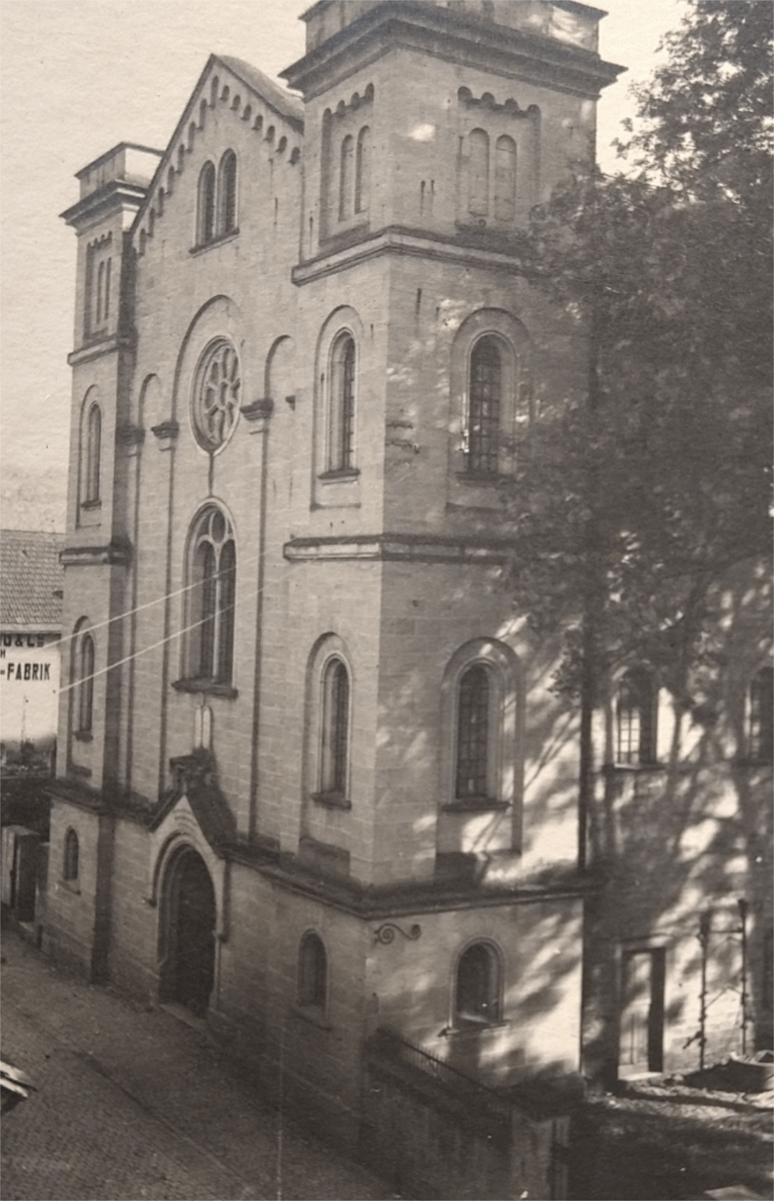 Historisches Gebäude mit neugotischer Architektur, vermutlich eine ehemalige Kirche oder Versammlungshaus, umgeben von Bäumen und einer gepflasterten Straße, im Hintergrund sichtbar eine Fabrik.