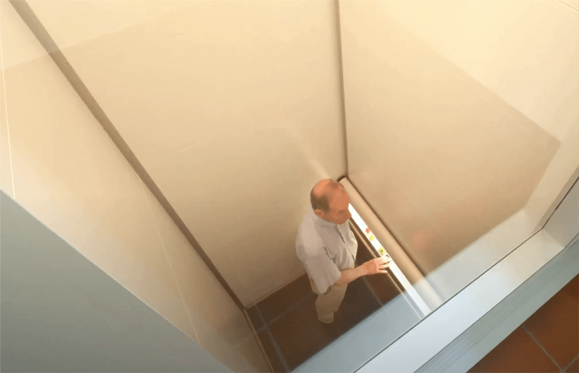 Ein älterer Mann steht in einem modernen Aufzug mit Glaswänden und schaut nach oben. Der Innenraum ist hell und minimalistisch gestaltet, was eine zeitgemäße Ästhetik vermittelt.