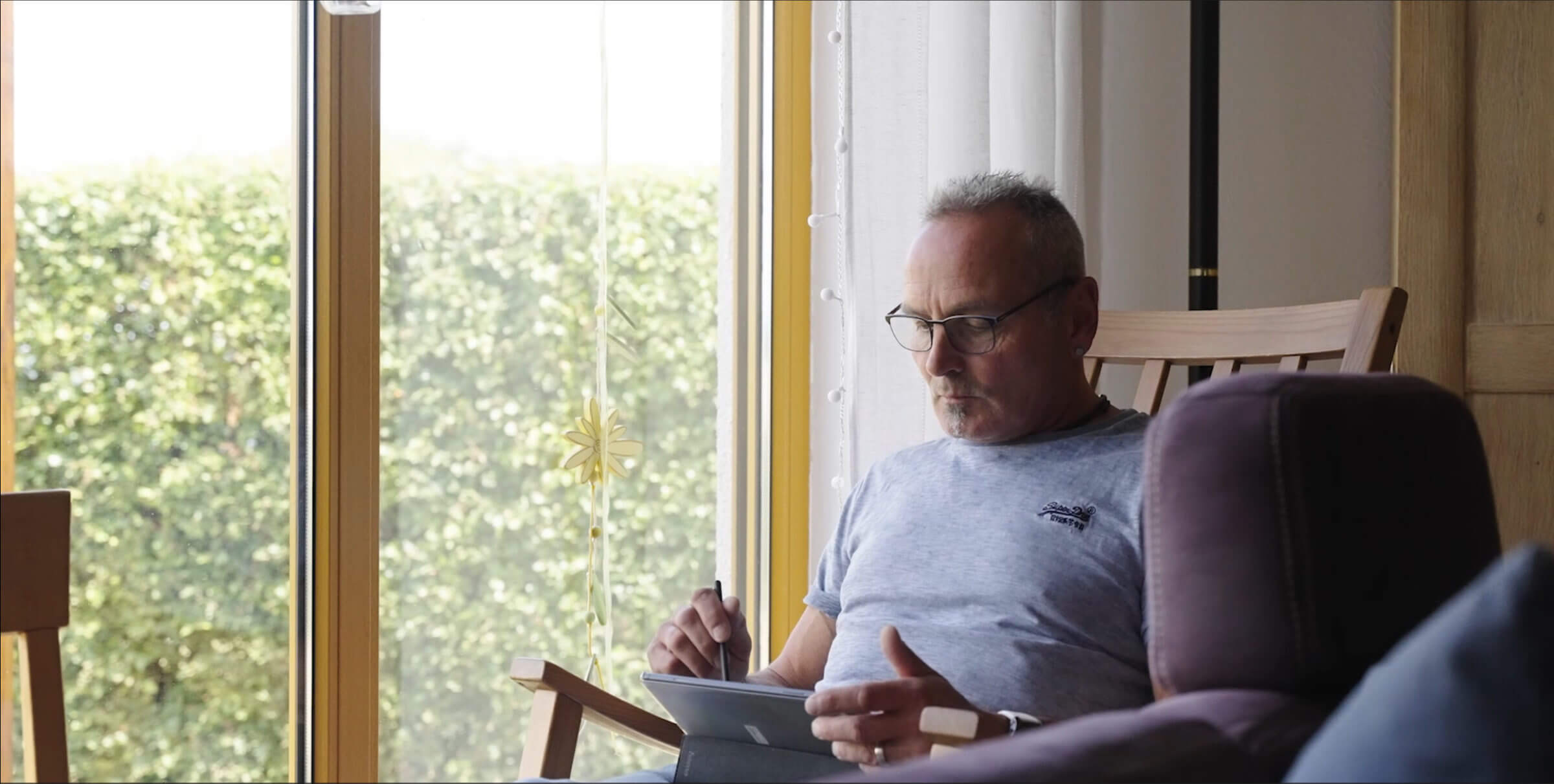 Älterer Mann sitzt entspannt in einem Sessel und liest auf einem Tablet, während er von natürlichem Licht durch große Fenster umgeben ist. Im Hintergrund ist eine grüne Hecke sichtbar, die eine ruhige Atmosphäre schafft.