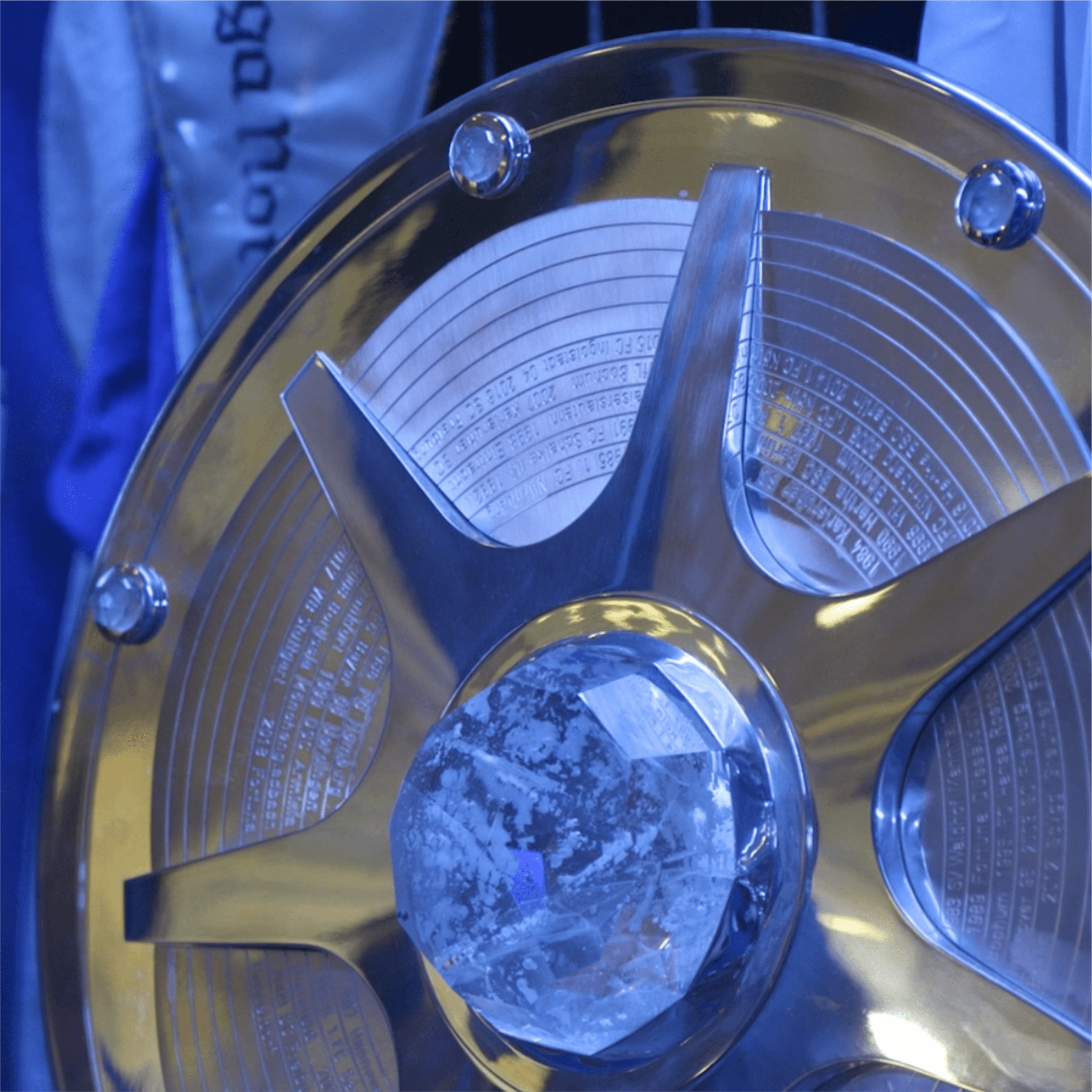 Meisterschale_2 Detailaufnahme eines glänzenden Pokals mit einer großen, klaren Kristallspitze und einem kunstvollen Design, umgeben von blauer Stoffverkleidung.