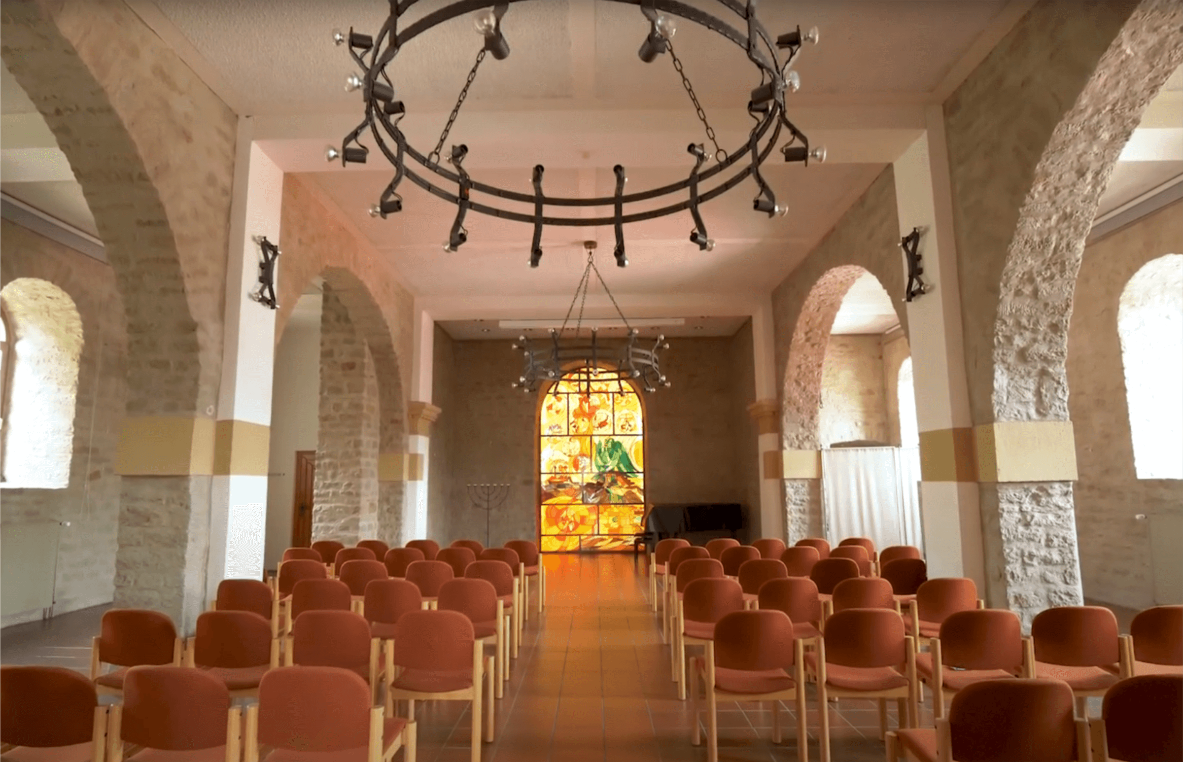 Innenansicht eines modernen Veranstaltungsraums mit Stühlen in orange, einem großen, bunten Glasfenster im Hintergrund und einer modernen Beleuchtung. Der Raum hat hohe, gewölbte Decken und Steinwände, die eine einladende Atmosphäre schaffen. Ideal für Konferenzen, Feiern oder kulturelle Veranstaltungen.