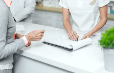 Frau in medizinischer Kleidung notiert Informationen am Empfangstresen, während eine andere Person Fragen stellt. Die Szene zeigt eine professionelle Atmosphäre in einer Praxis oder Klinik, die auf Patientenkontakt und Service ausgerichtet ist.