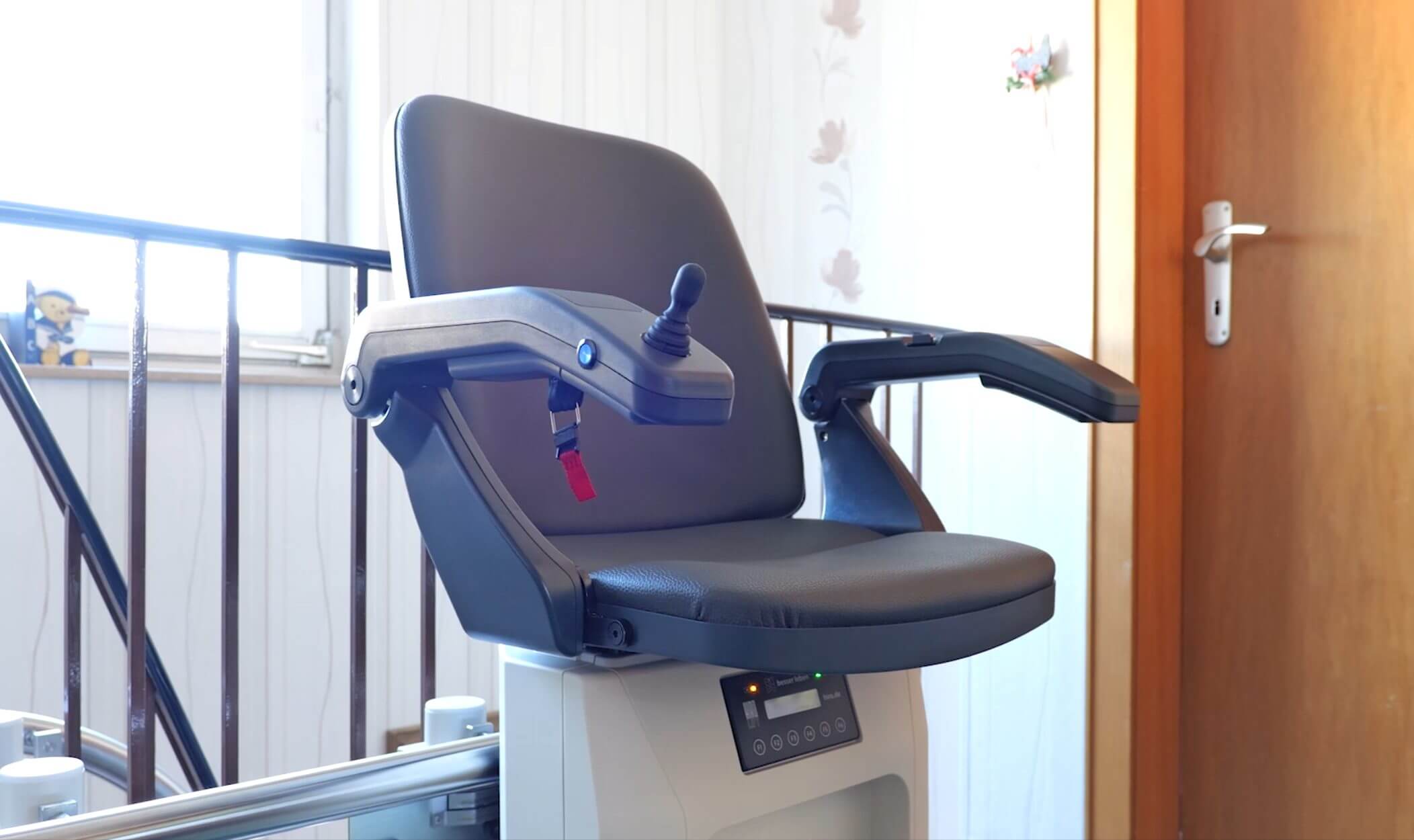 Bequemer Treppenliftstuhl mit Armlehnen und Steuerhebel, ideal für barrierefreies Wohnen, in einem hellen Raum mit Holztür im Hintergrund.