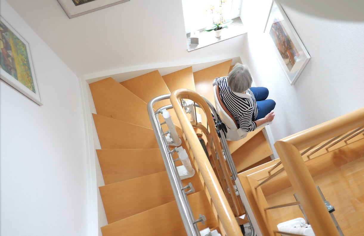 Eine Person sitzt auf einer Treppe in einem hellen Treppenhaus mit Holzstufen und einem Handlauf. An den Wänden hängen Bilder, und ein Fenster lässt Tageslicht herein.