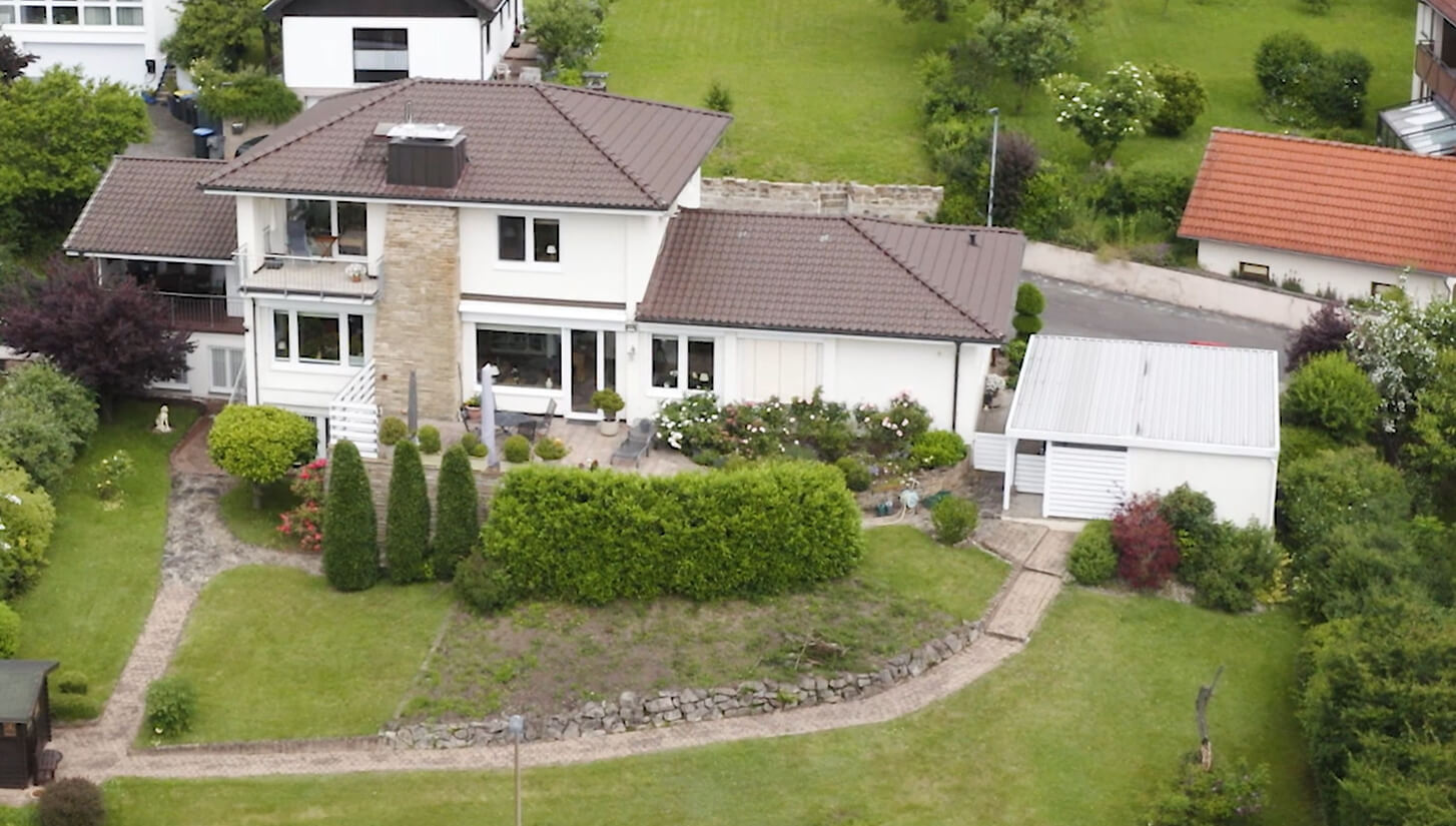 Luftaufnahme eines modernen Einfamilienhauses mit gepflegtem Garten, umgeben von grünen Pflanzen und Bäumen. Die Terrasse ist mit Blumen dekoriert, und ein kleiner Gartenweg führt zur Garage.
