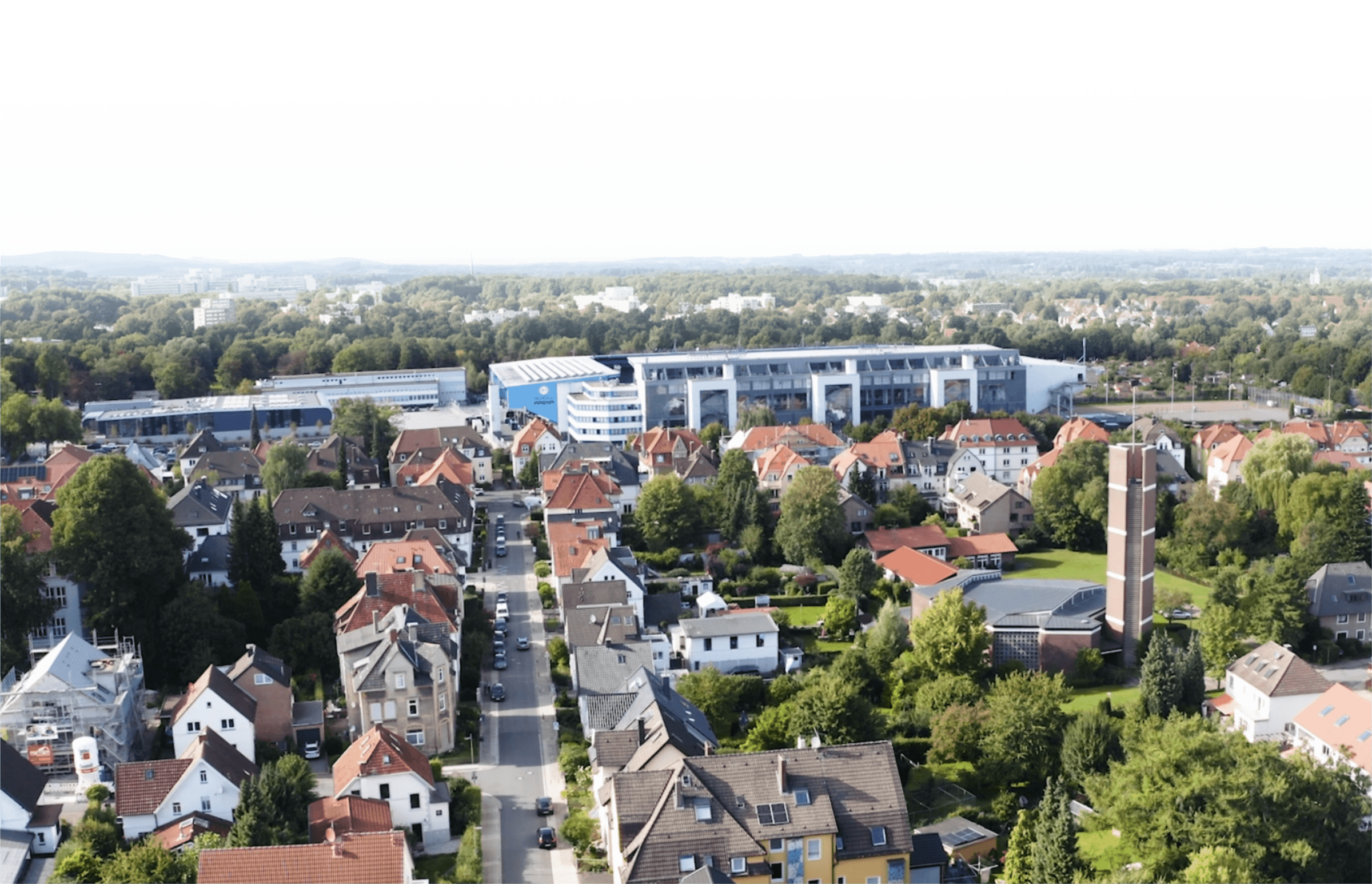 Stadion_Teuto Luftaufnahme einer Wohngegend mit Einfamilienhäusern, grünen Gärten und einem modernen Gebäude im Hintergrund, umgeben von Bäumen und einer weitläufigen Landschaft.