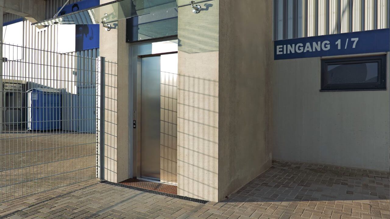 Eingang zu einem modernen Gebäude mit Glasfront und Metallzaun, beschildert mit "EINGANG 1/7", umgeben von gepflastertem Boden und industrieller Umgebung.