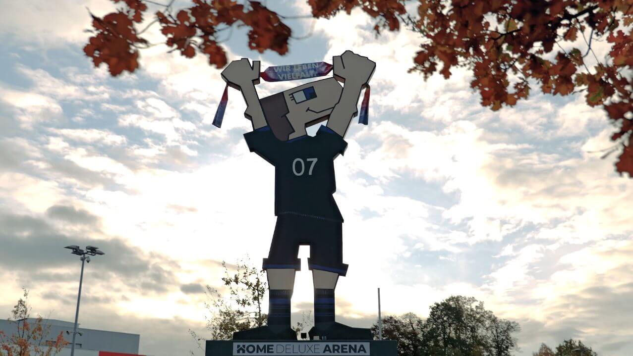 Große Statue eines Fußballspielers mit der Nummer 07, die in der Heimat-Arena steht, umgeben von herbstlichen Bäumen und einem bewölkten Himmel.