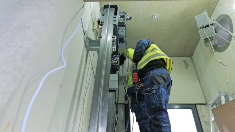 Techniker bei der Installation eines Aufzugsystems in einem modernen Gebäude, ausgestattet mit Sicherheitsweste und Werkzeug, während er an der mechanischen Struktur arbeitet.