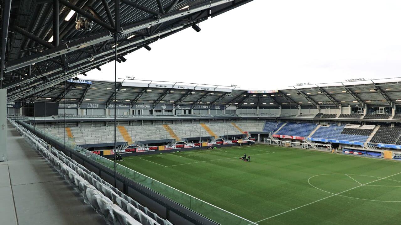 Blick auf das Innere eines modernen Fußballstadions mit grüner Spielfläche, leeren Zuschauerplätzen und überdachtem Tribünenbereich. Ideal für Sportveranstaltungen und große Events.