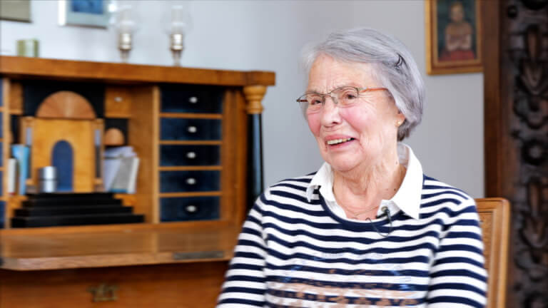 Ältere Frau mit Brille und grau meliertem Haar, die in einem gemütlichen Raum sitzt. Sie trägt ein gestreiftes Oberteil und lächelt freundlich. Im Hintergrund ist ein Holzschrank mit verschiedenen Gegenständen zu sehen.