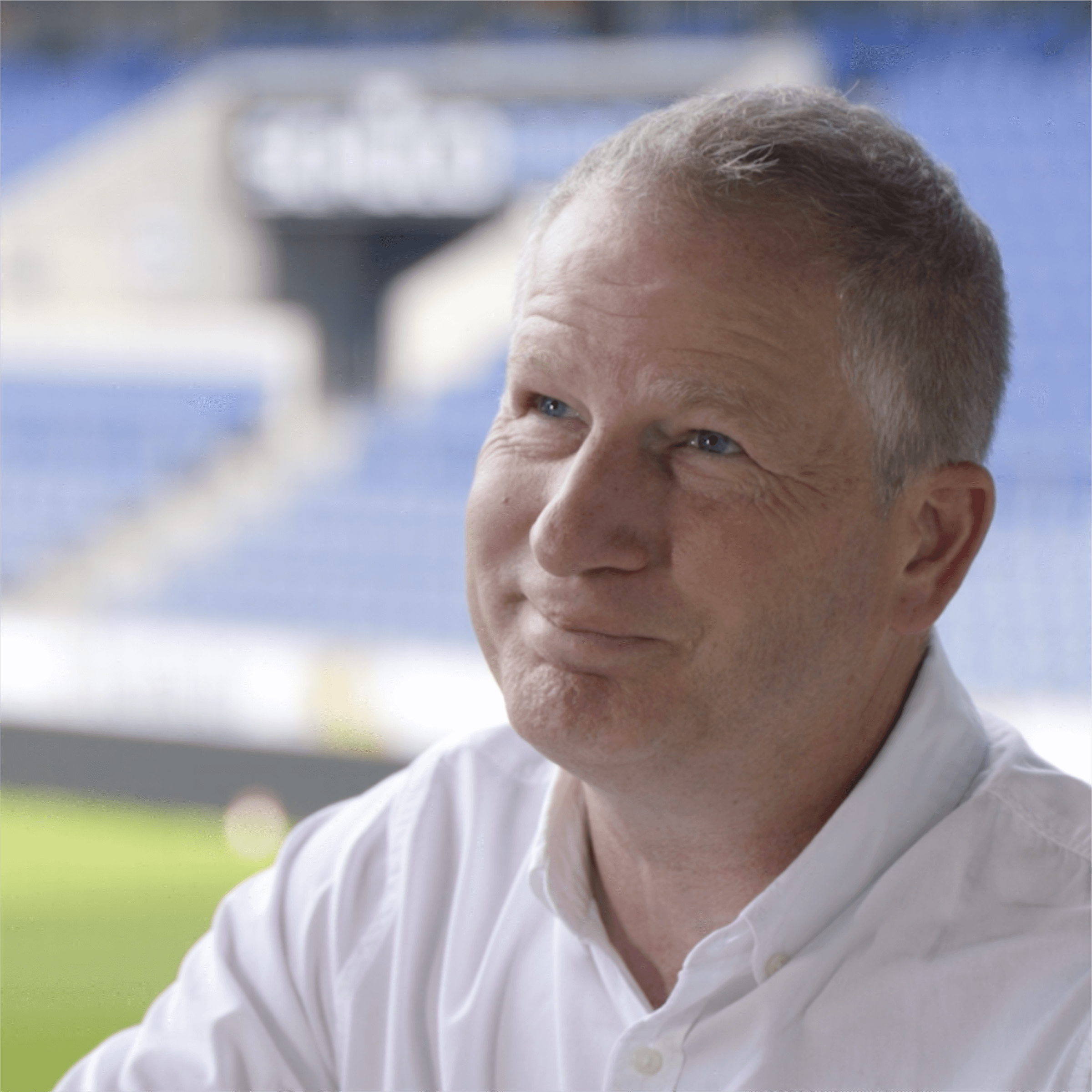 Venghaus Mann in einem Stadion mit nachdenklichem Ausdruck, trägt ein weißes Hemd und sitzt vor einer leeren Tribüne.