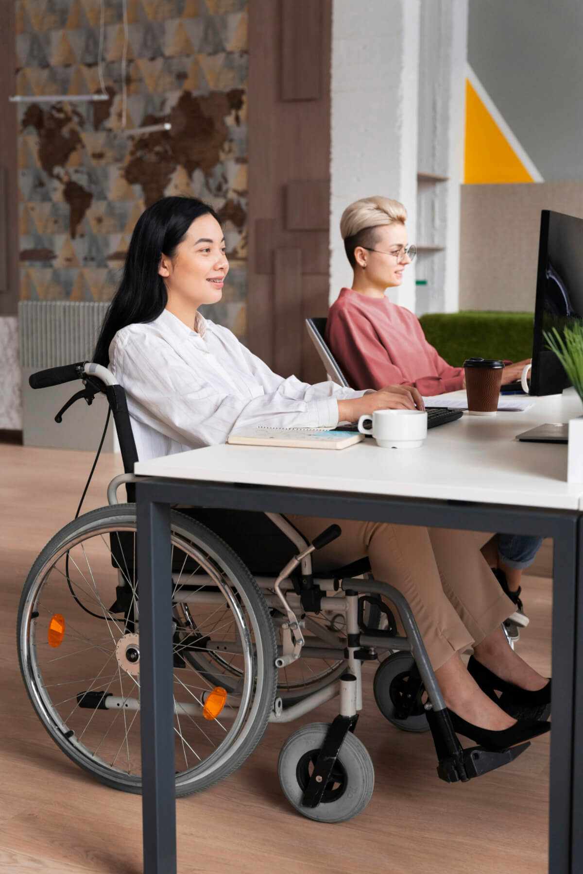 Zwei Frauen arbeiten in einem modernen Büro, eine davon sitzt im Rollstuhl. Beide konzentrieren sich auf ihre Computerbildschirme, während eine Tasse Kaffee und ein Notizbuch auf dem Tisch stehen. Der Raum ist hell und einladend gestaltet.